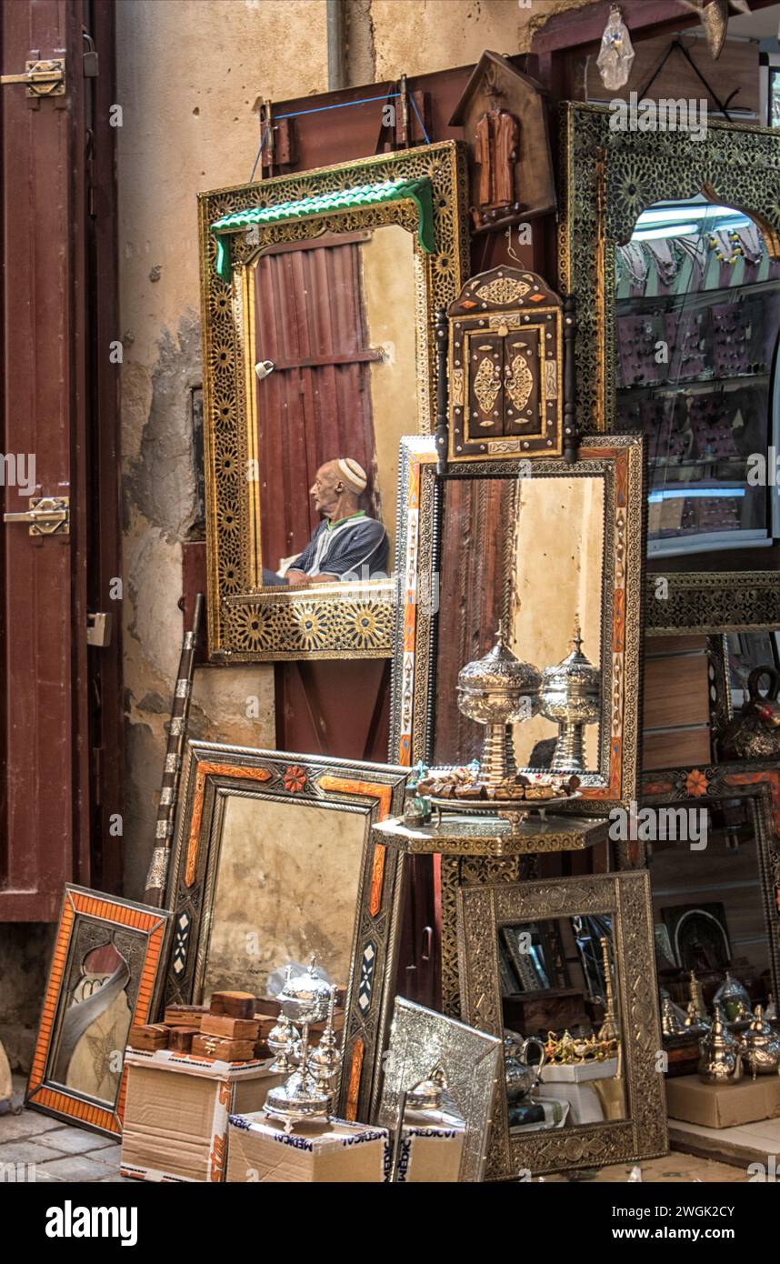 Spiegelbild eines Händlers im Spiegel eines Ladens in der Medina von Fès. Fès, die zweitgrößte Stadt Marokkos, wird oft als Kulturhauptstadt des Landes bezeichnet. Es ist vor allem bekannt für seine von Fes El Bali ummauerte Medina mit mittelalterlicher marinidischer Architektur, pulsierenden Souks und der Atmosphäre der alten Welt. Die Medina beherbergt religiöse Schulen wie die Bou Inania aus dem 14. Jahrhundert und Al Attarine, die beide mit aufwändigen Zedernschnitzereien und kunstvollen Fliesen verziert sind. Marokko. Stockfoto