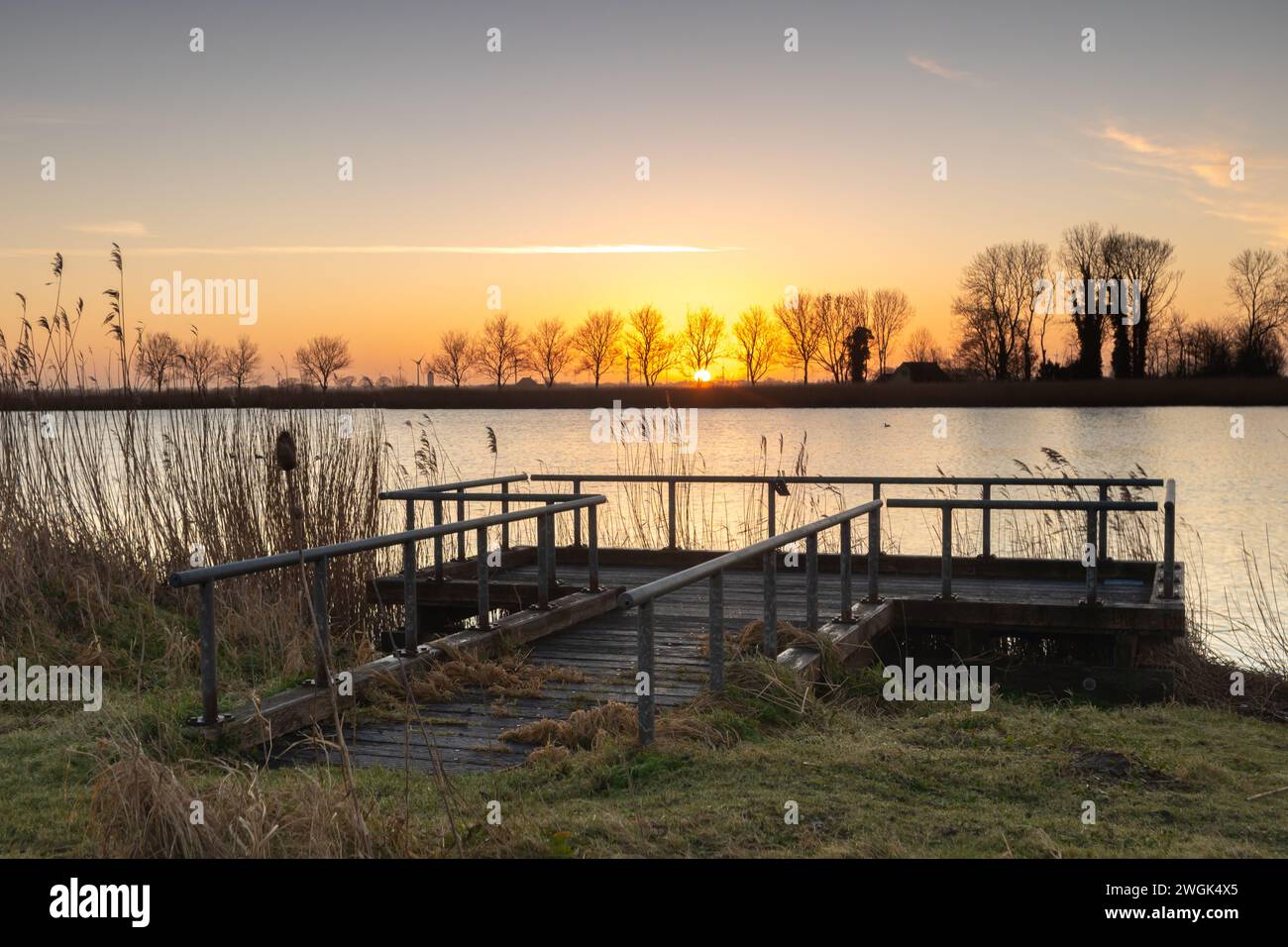 Der Steg scheint verlassen zu sein und wartet vielleicht auf ein paar Fischer. Das Schilf an der Seite und die Bäume im Hintergrund verleihen diesem Foto eine ländliche Atmosphäre Stockfoto