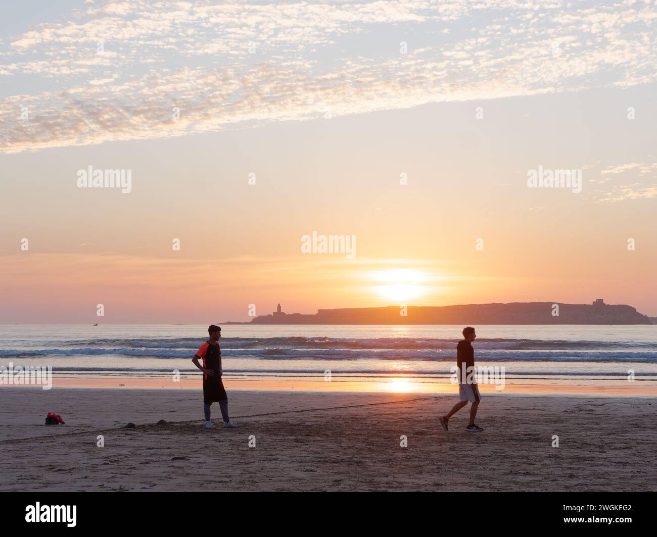 Junge Männer in Silhouette spielen Fußball an einem Sandstrand bei Sonnenuntergang mit einer Insel dahinter in Essaouira, Marokko, 5. Februar 2024 Stockfoto