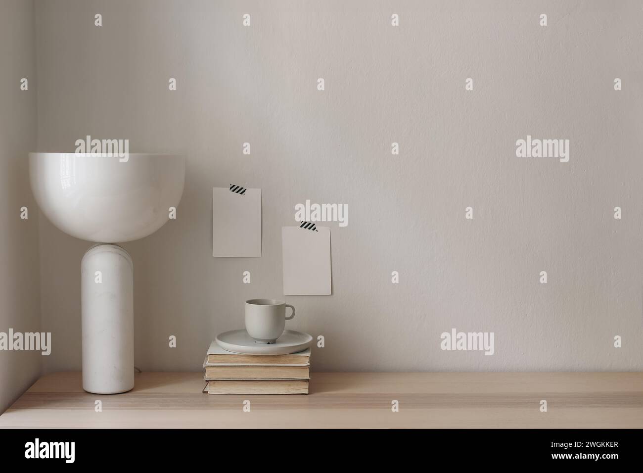 Leere Grußkarten Mockups, die auf weiße Wand aufgeklebt sind. Elegante moderne Marmorlampe, geometrischer Schatten. Holztisch, Schreibtisch mit alten Büchern und einer Tasse davon Stockfoto