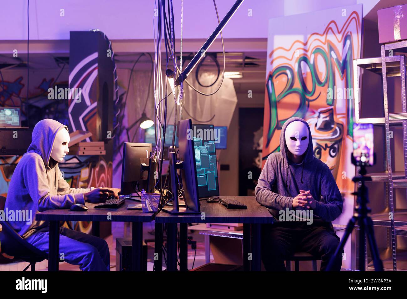 Internetbetrüger in anonymen Masken bedrohen Opfer online auf dem Smartphone. Hacker, die während ihrer Zusammenarbeit in einem verlassenen Lager Videos von Lösegeldnachrichten auf dem Mobiltelefon aufzeichnen Stockfoto
