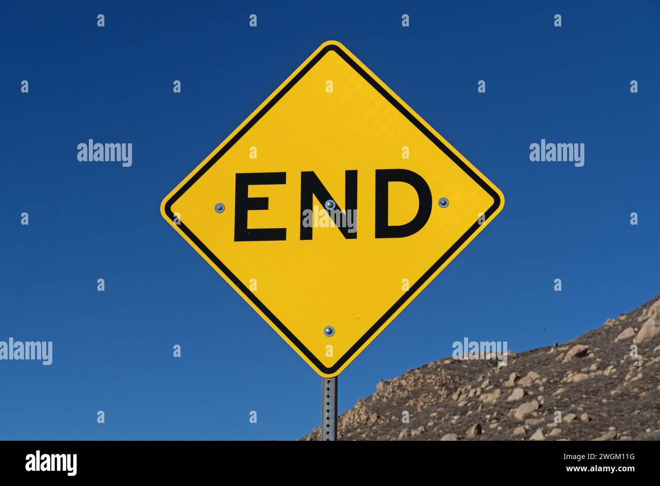 Gelbes und schwarzes Straßenschild mit blauem Himmel und felsigem Hügel dahinter Stockfoto