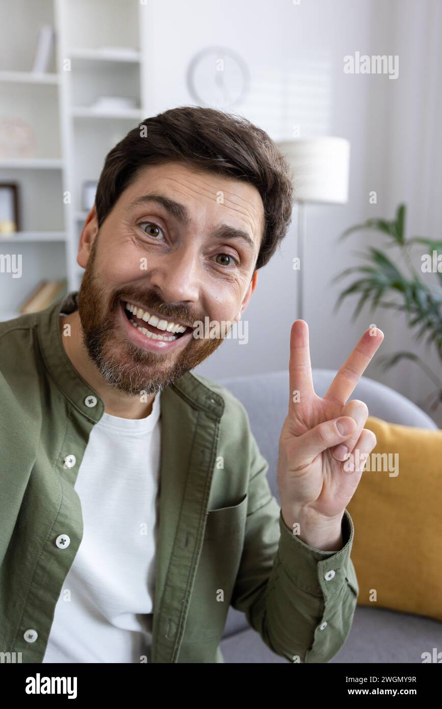 Porträt eines glücklichen bärtigen Mannes, der ein Friedenszeichen mit zwei Fingern über dem Hintergrund des Wohnhauses macht. Gutaussehender Kerl, der Selfie auf der Frontkamera des Smartphones macht oder Videoanrufe startet und einen Freund begrüßt. Stockfoto