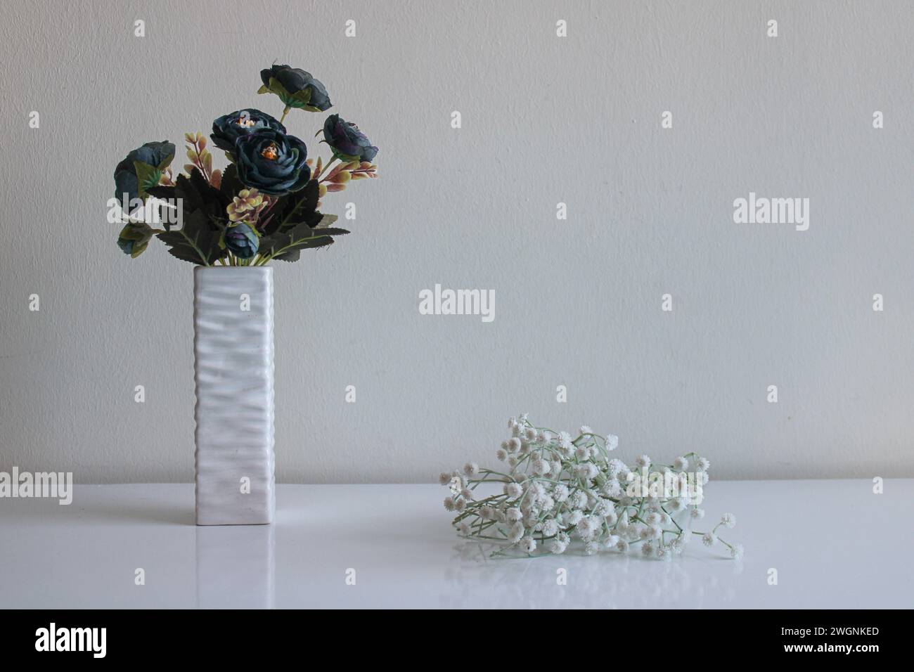 Ein Blumenstrauß aus künstlichen blauen Blumen steht in einer Relief-Vase. Kleine weiße Blumen liegen in der Nähe. Neutraler Hintergrund und Oberfläche Stockfoto