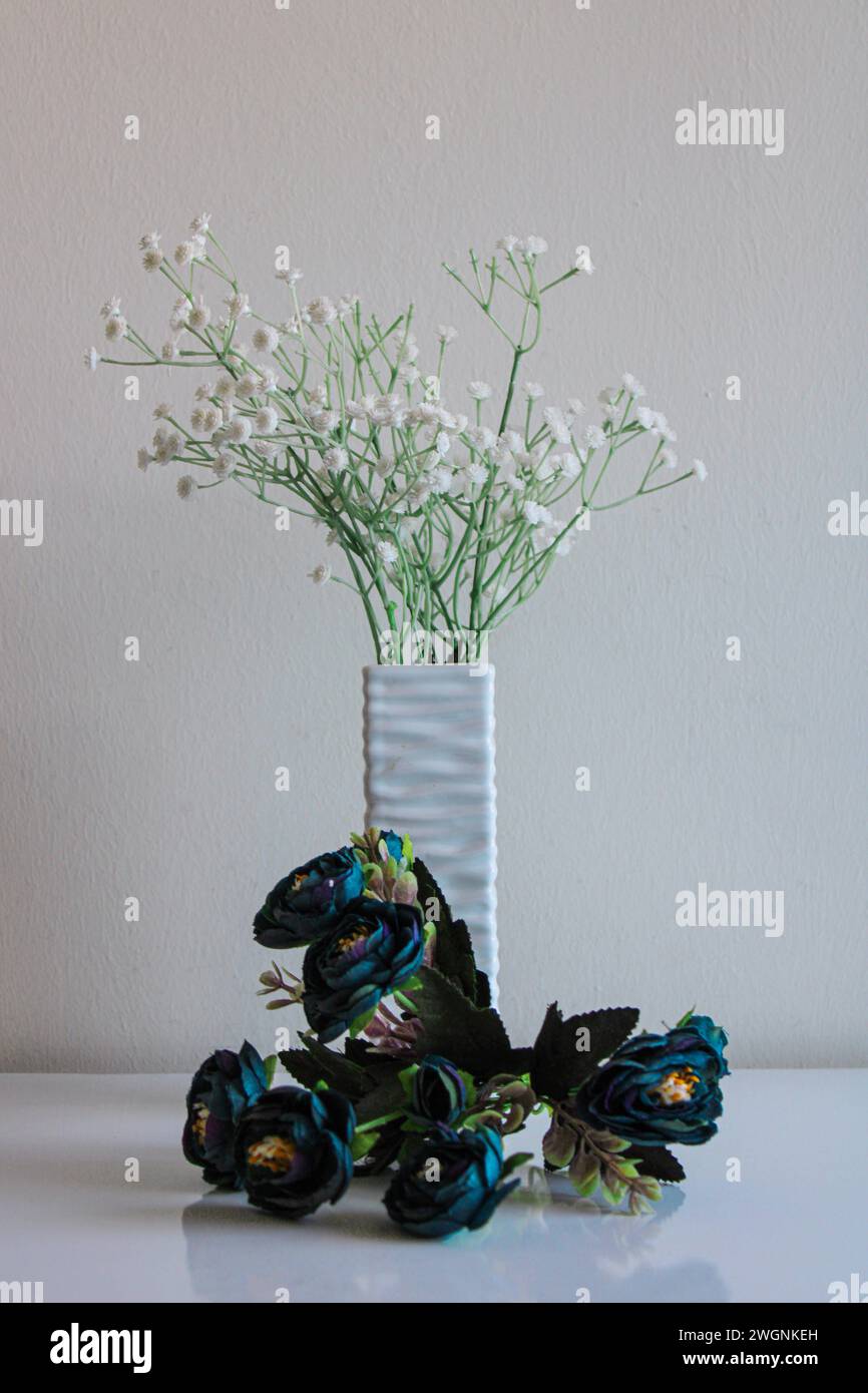 Eine Vase mit Relief-Textur steht auf einer weißen Oberfläche vor einem neutralen Hintergrund. Weiße und blaue Kunstblumen in Blumensträußen Stockfoto