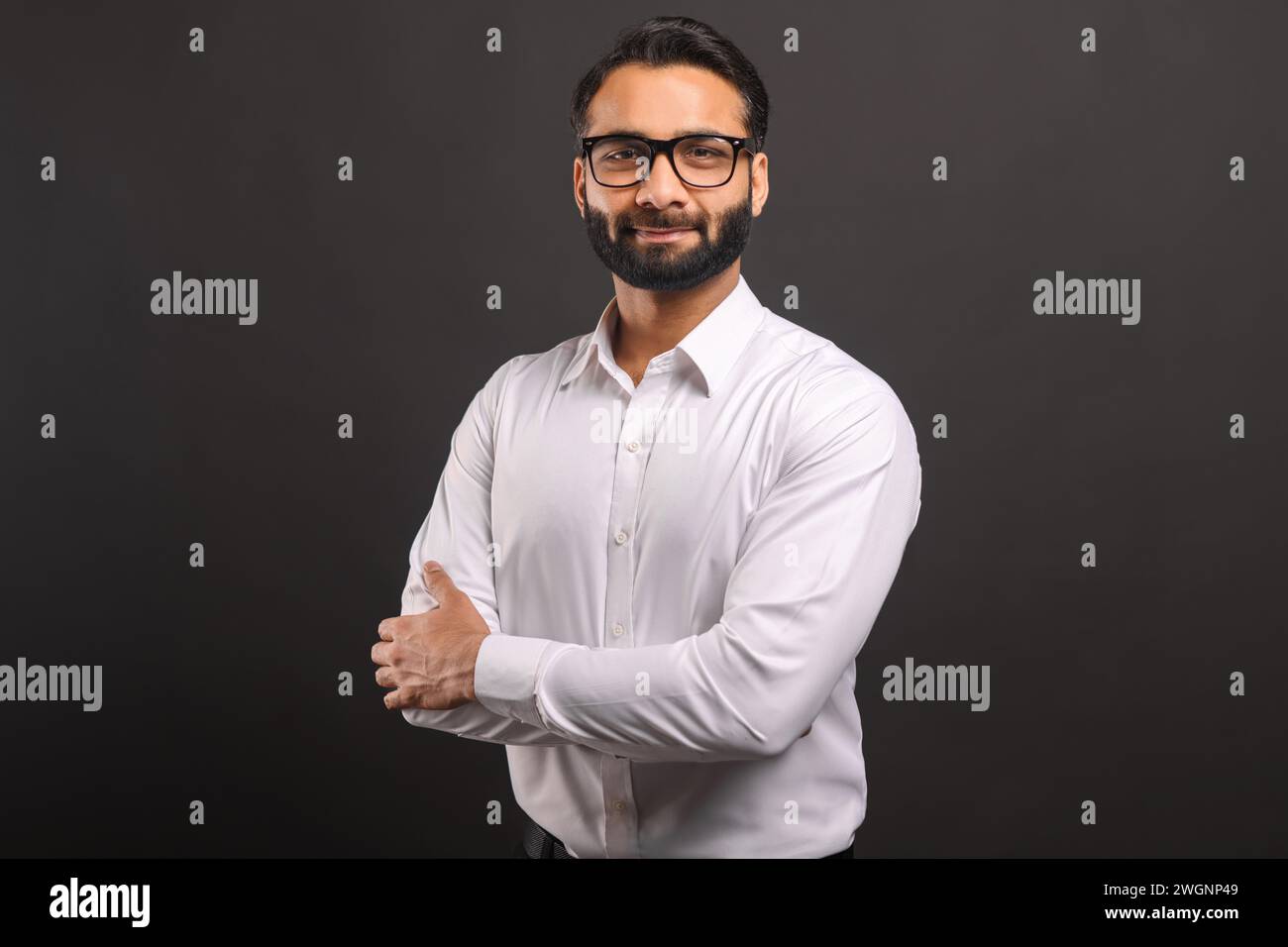 Kopfaufnahme eines intelligenten, professionellen indischen Geschäftsmannes mit formeller Kleidung und Brille, der auf die Kamera blickt, während er die Arme getrennt auf Schwarz posiert Stockfoto