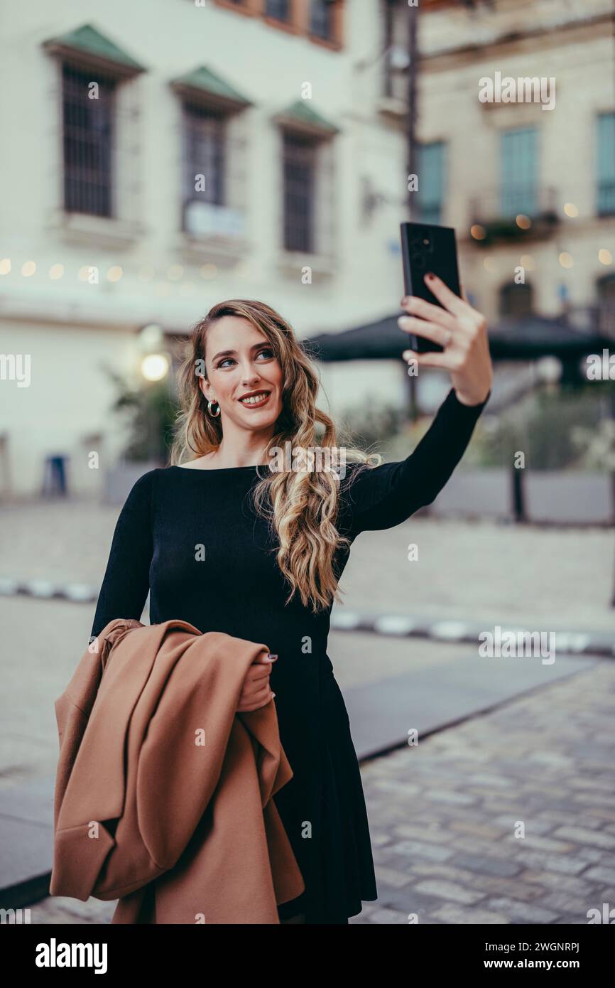 Eine modische Kaukasierin, die ein Selfie auf einer Stadtstraße macht, in einem schwarzen Kleid Stockfoto