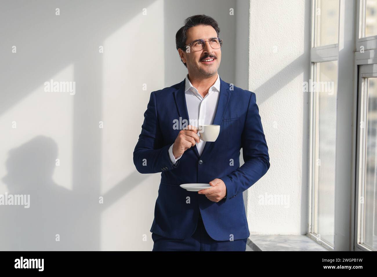 Lächelnder reifer Geschäftsmann in einem scharfen blauen Anzug, der eine Tasse Kaffee genießt, während er aus dem Fenster blickt Stockfoto