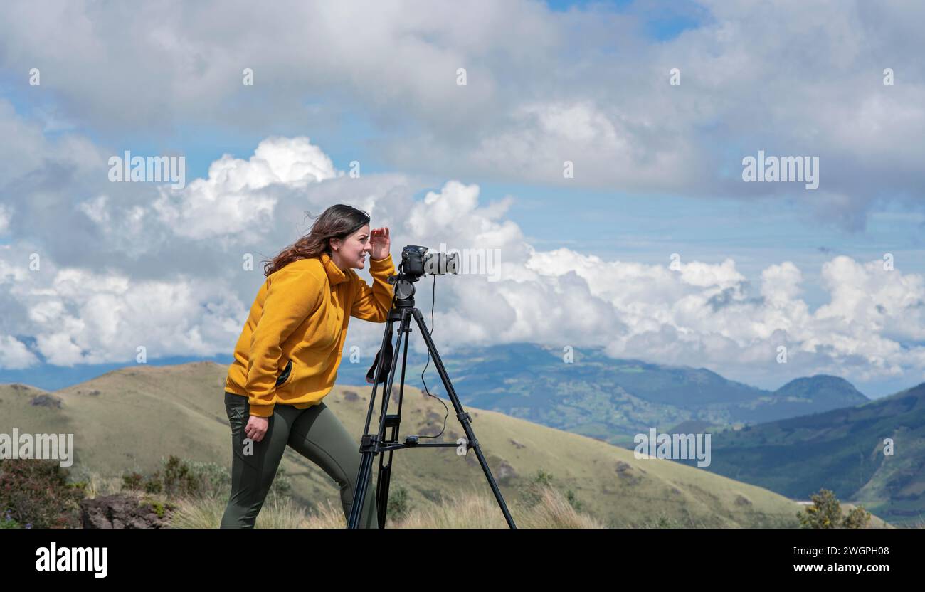 Schöne junge lateinamerikanische Frau mit Kamera und Stativ, die an einem sonnigen Tag Fotos von Landschaften auf dem Gipfel eines Berges macht Stockfoto