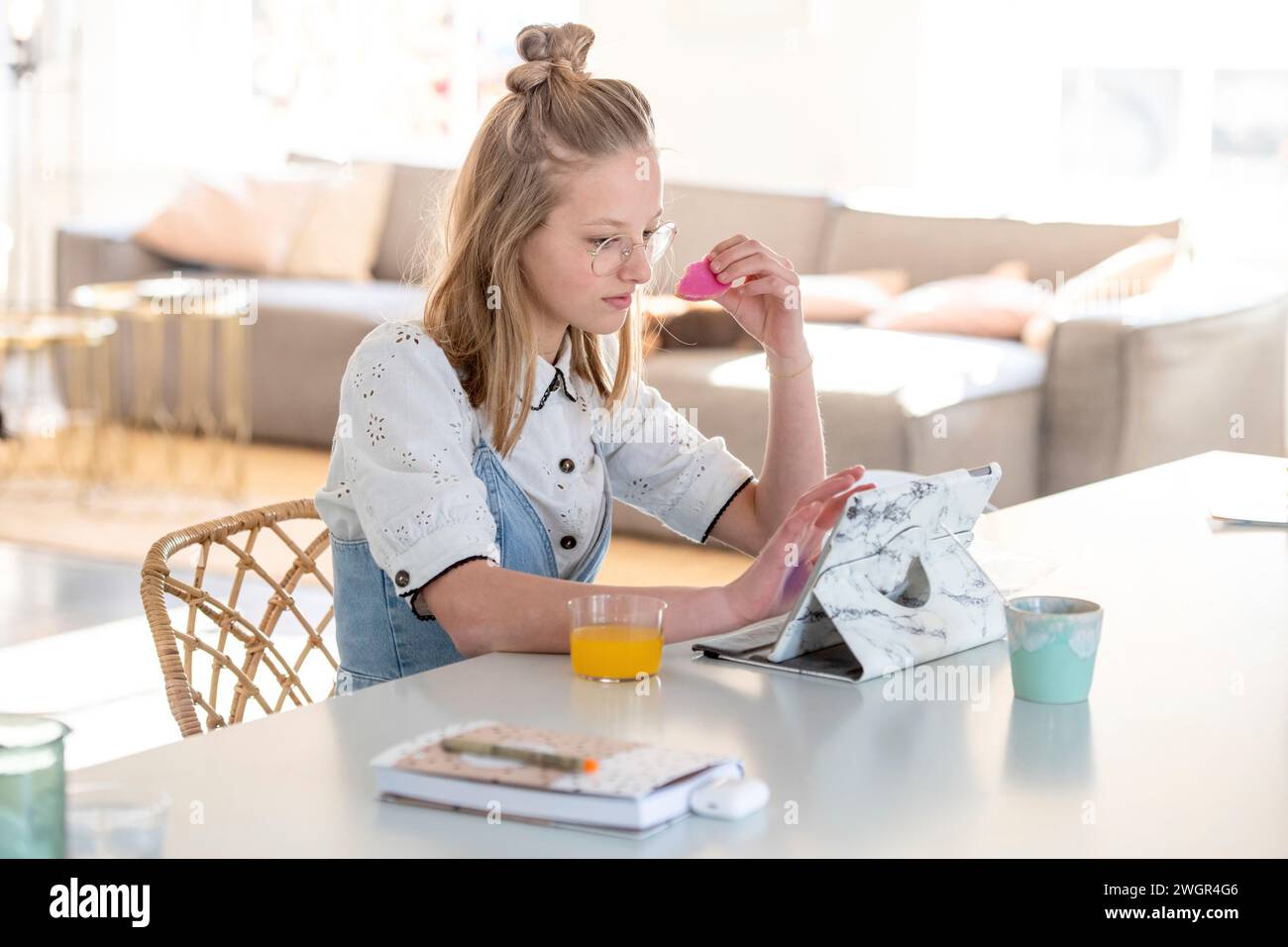 Die blonde attraktive junge Frau sitzt am Küchentisch und isst beim Stöbern auf ihrem Tablet Stockfoto
