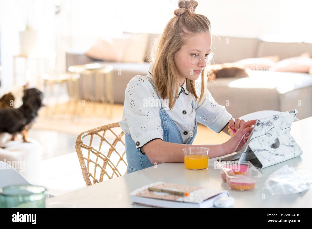 Die blonde attraktive junge Frau sitzt am Küchentisch und isst beim Stöbern auf ihrem Tablet Stockfoto