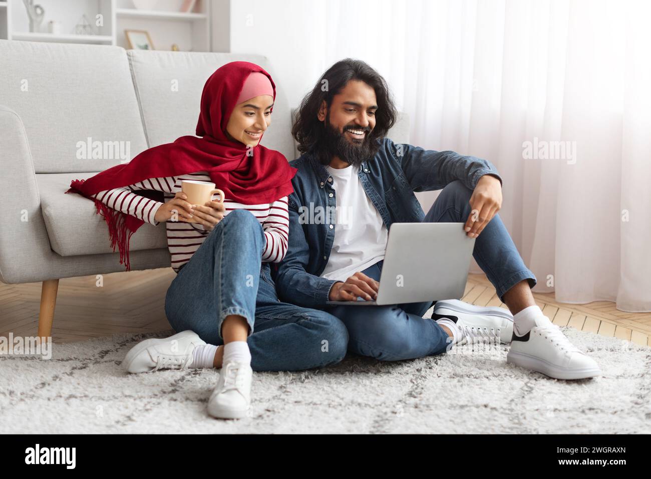 Entspanntes, junges muslimisches Paar, das mit Laptop und Kaffeetasse auf dem Boden sitzt Stockfoto