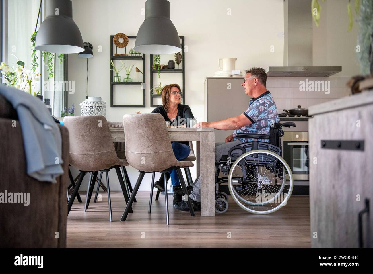 Die Frau bereitet ihr Mittagessen vor, der in seinem Rollstuhl am Tisch sitzt Stockfoto