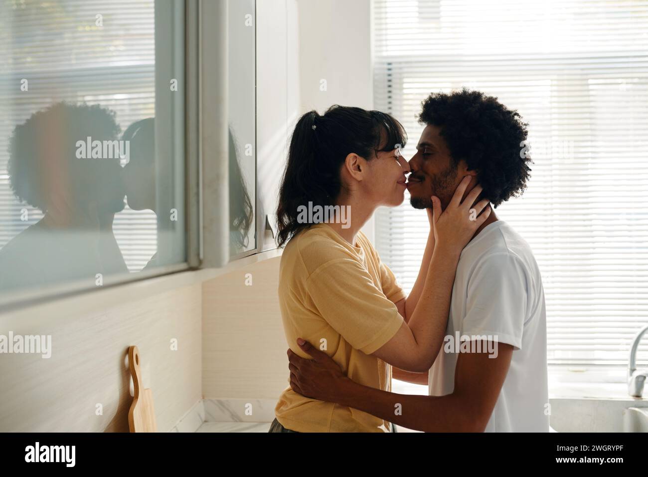 Junge, liebevolle interkulturelle Ehemann und Ehefrau in Casualwear, die sich küssen, während die Frau die Hände des Mannes am Hals hält Stockfoto