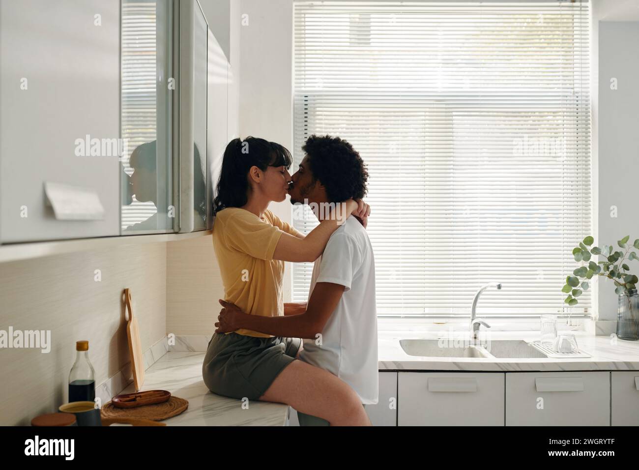 Junge, liebevolle Frau in Casualwear, die auf der Küchentheke sitzt und ihren Mann küsst und die Hände um ihre Taille hält Stockfoto