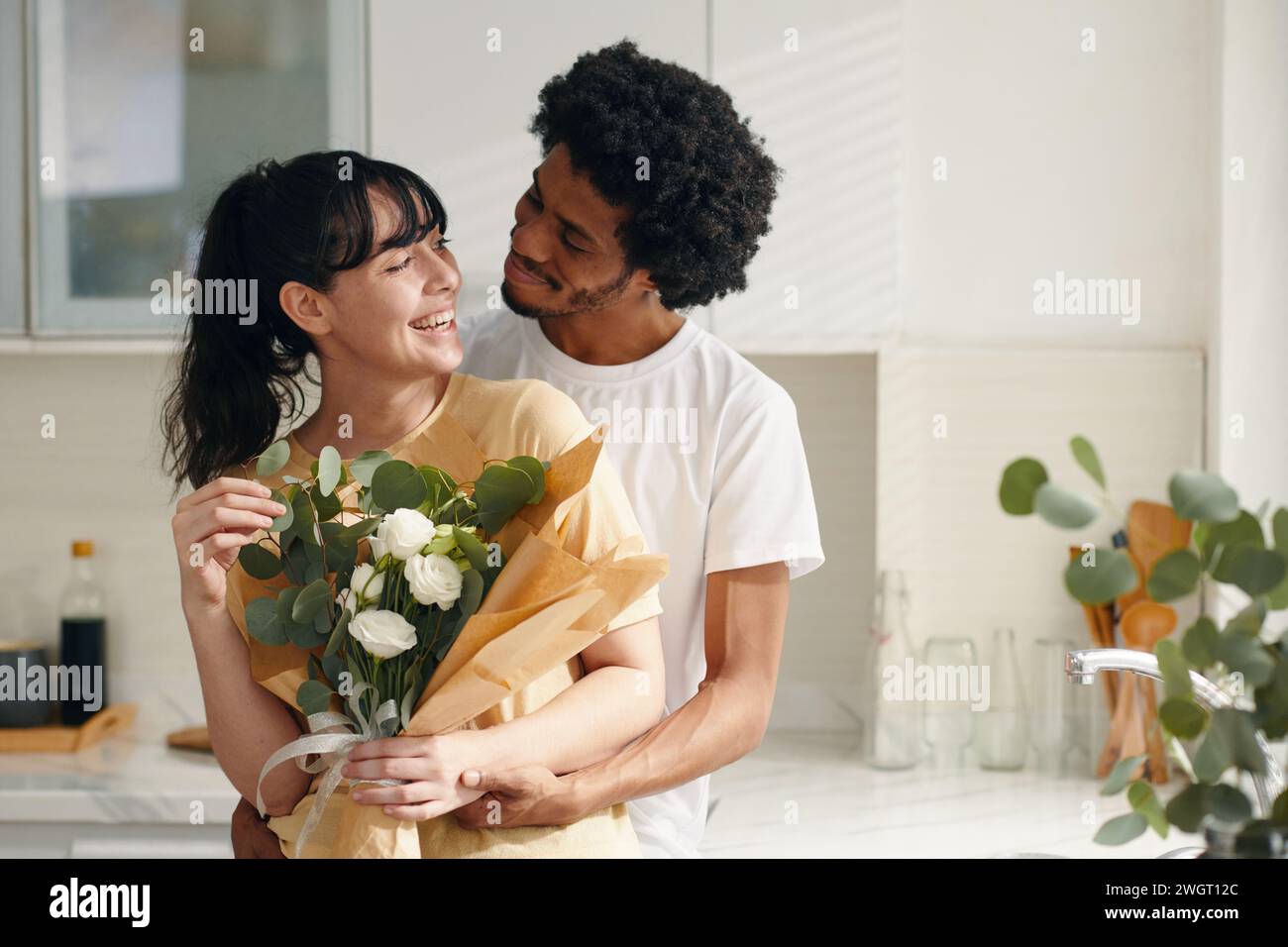 Ein junges, liebevolles Paar in Casualwear, das sich ansieht, während der Kerl seine Freundin umschließt, die einen Haufen weißer Blumen hält Stockfoto