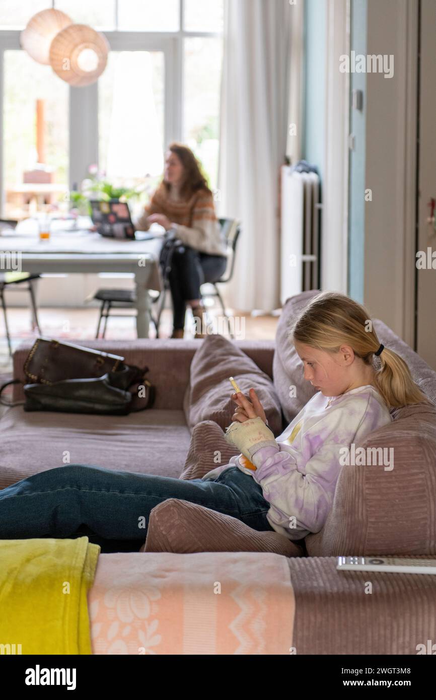Junge Teenagerin, die sich auf dem Sofa entspannt und mit ihrem Telefon recherchiert. Mit Großmutter und Mutter im Hintergrund, die sich eine Tasse Tee teilen Stockfoto