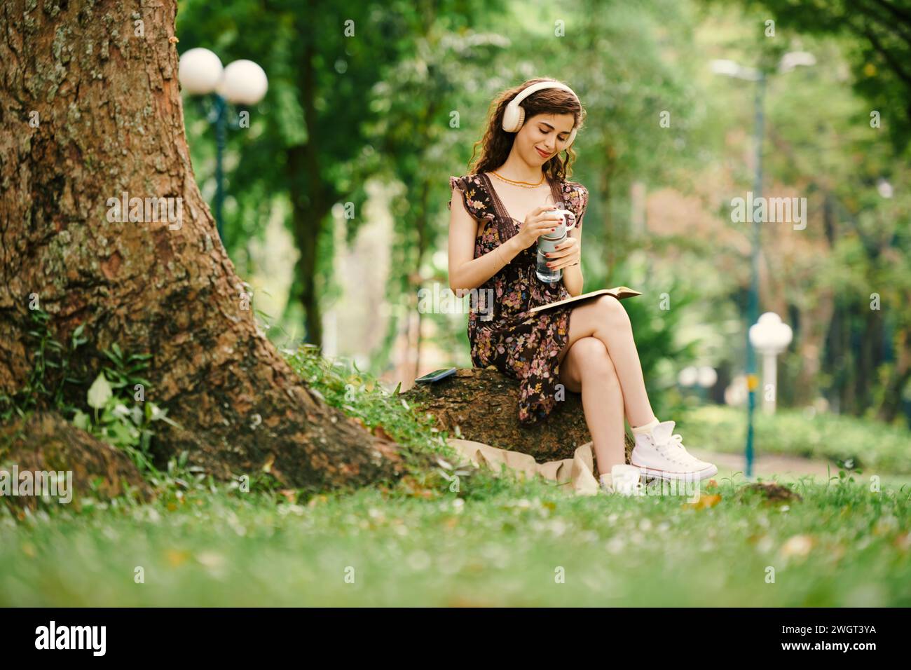 Lächelnde junge Frau in Freizeitkleidung und Kopfhörern, die im Park unter einem Baum sitzt, Musik hört und Märchenbücher oder Tagebücher liest Stockfoto