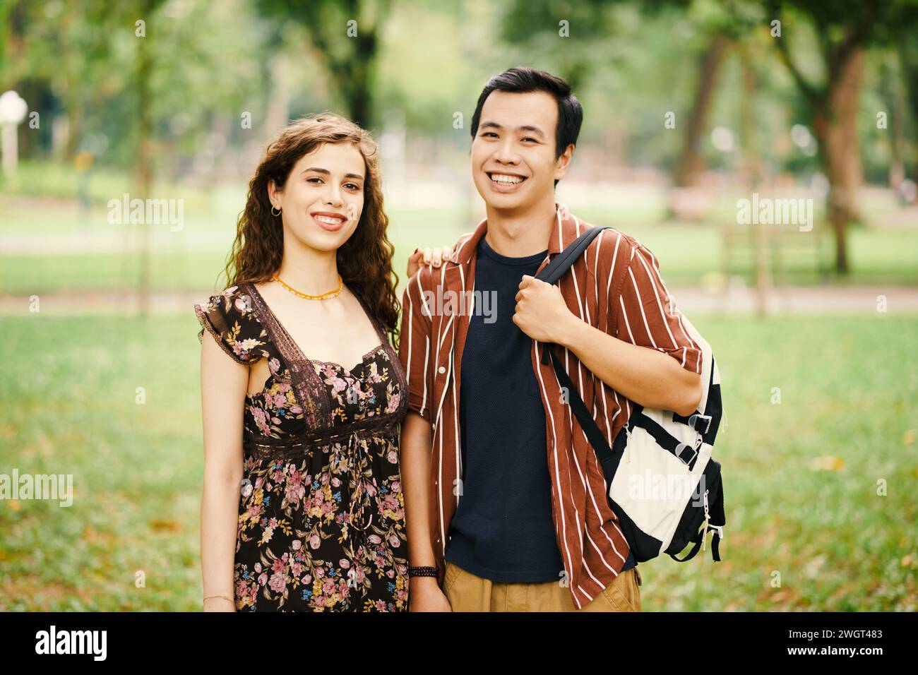 Glückliches junges interkulturelles Paar in Casualwear, das im Sommer auf dem Campus oder im großen Park mit einem Lächeln in die Kamera blickt Stockfoto