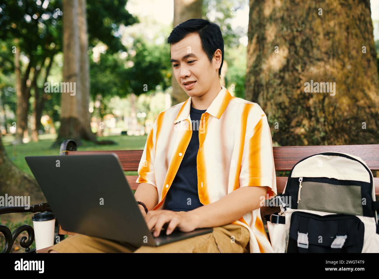 Junger asiatischer Geschäftsmann in Casualwear, der auf den Laptop-Bildschirm schaut und auf einer Bank im Park mit vielen Bäumen schreibt Stockfoto