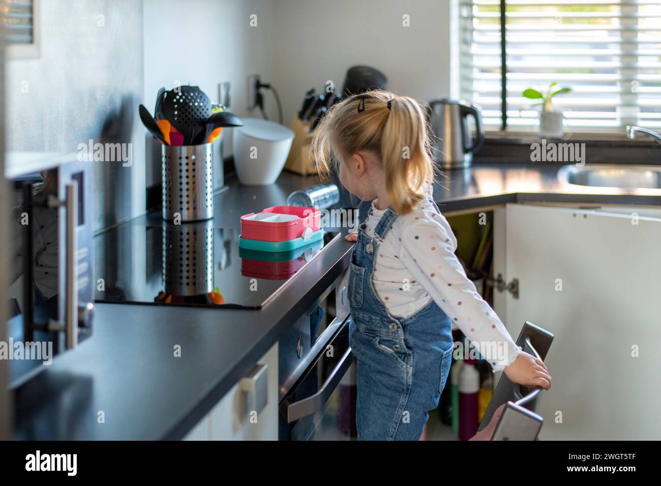 Blondes kleines Mädchen, das auf einem Stuhl in der Küche steht und darauf wartet, dass ihre Mutter ihr das Kochen beibringt Stockfoto