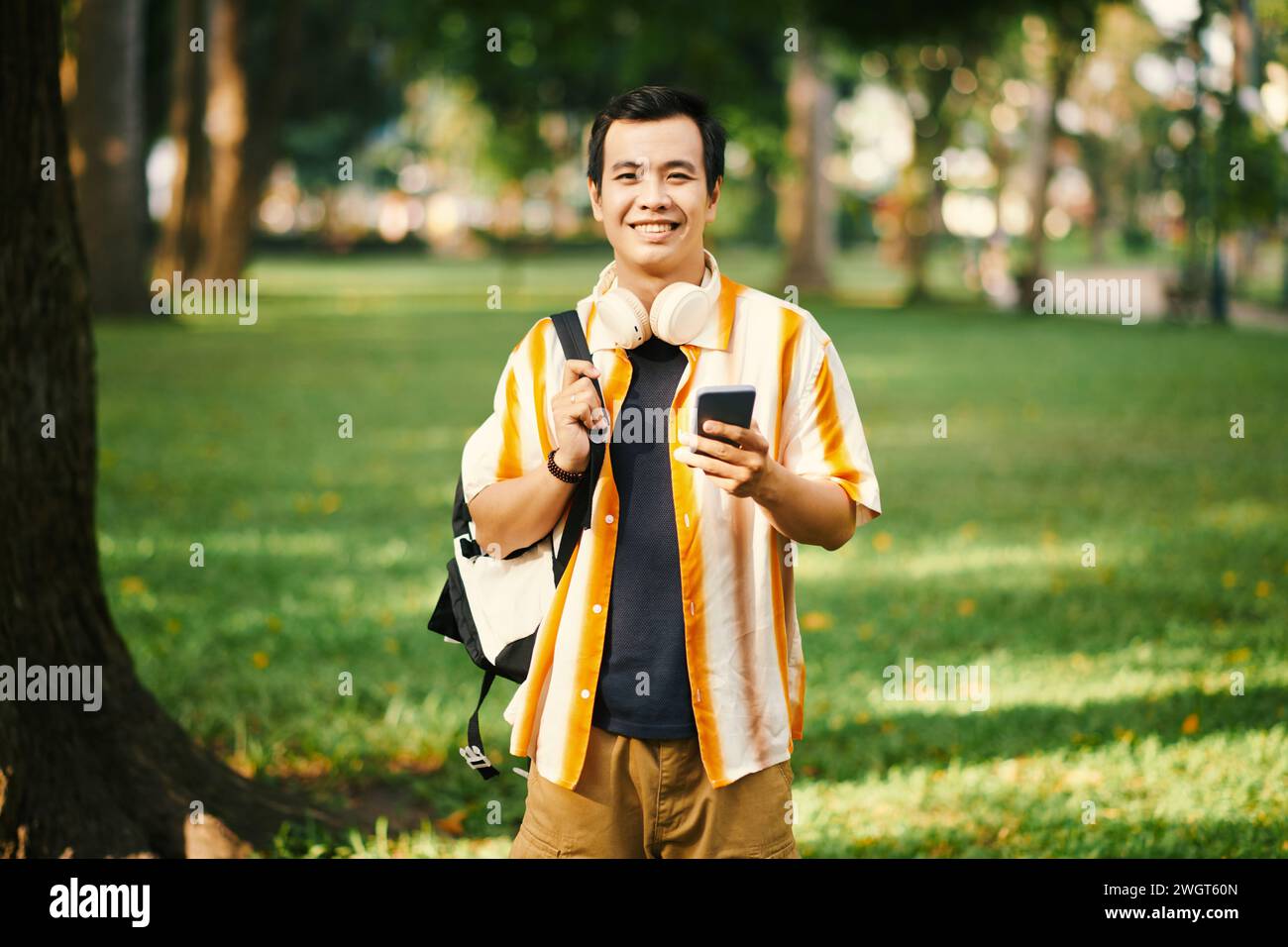 Junger, fröhlicher Mann in Casualwear, der Rucksack auf der Schulter und Smartphone in der Hand hält, während er im Park mit grünen Rasenflächen steht Stockfoto