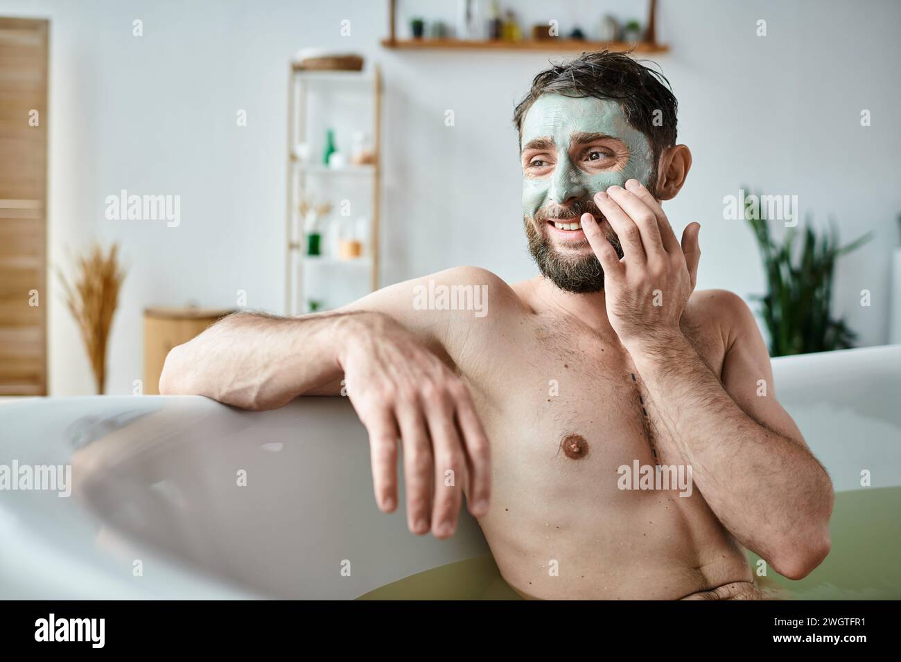 Fröhlicher gutaussehender Mann mit Bart und Gesichtsmaske, der in seiner Badewanne chillt, geistiges Gesundheitsbewusstsein Stockfoto