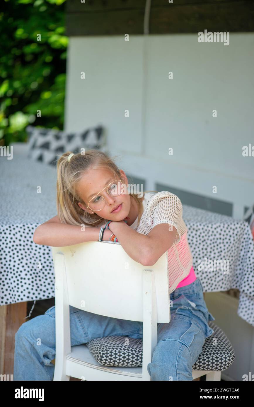 Blondes Teenagermädchen sitzt zu Hause auf einem Stuhl und genießt die Sonne und blickt in Ruhe und glücklich in die Kamera Stockfoto