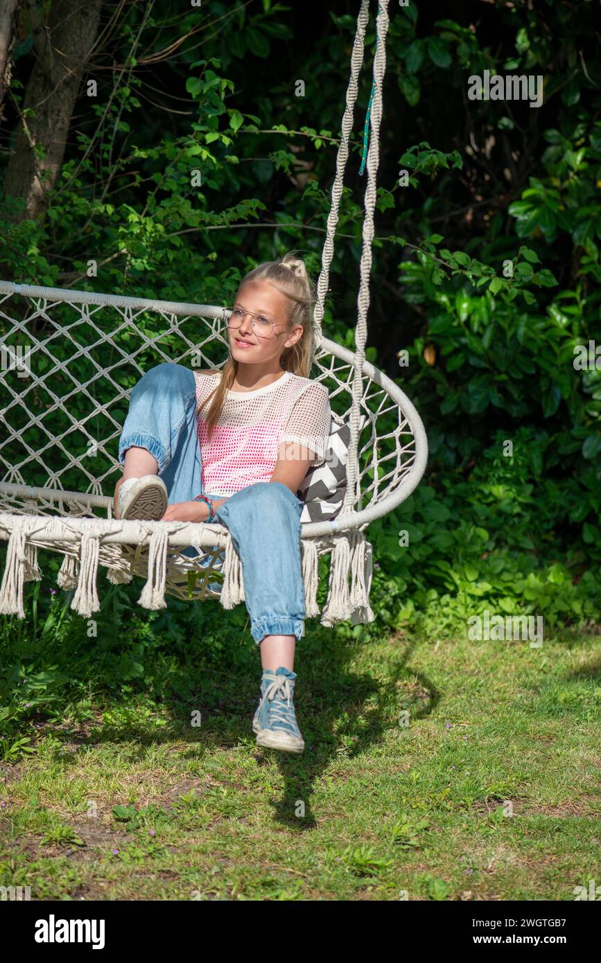 Blondes Teenagermädchen sitzt zu Hause auf einem Stuhl und genießt die Sonne und blickt in Ruhe und glücklich in die Kamera Stockfoto
