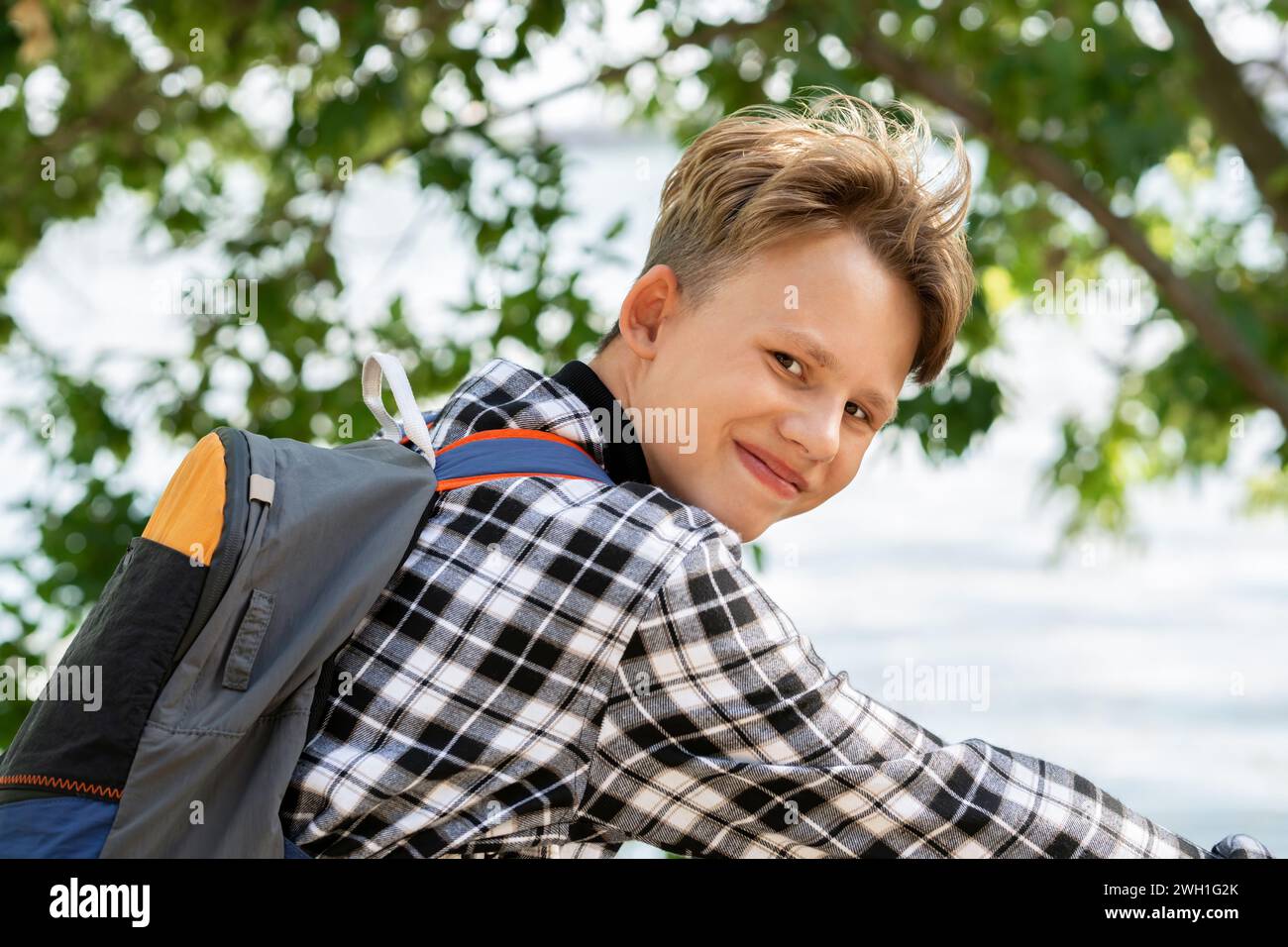 Porträt eines lächelnden Jungen mit blonden Haaren im karierten Hemd mit Rucksack, der auf dem Fahrrad sitzt, auf verschwommenen Bäumen und reflektierendem Teichhintergrund am sonnigen Tag Stockfoto