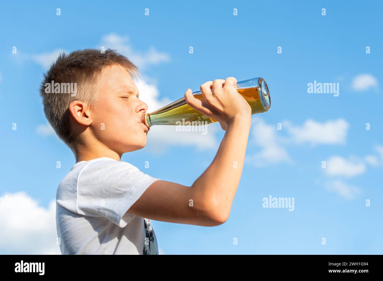 Fröhlicher Junge in weißem T-Shirt, der Limonade aus einer Glasflasche vor einer wunderschönen Himmelskulisse trinkt. Seitenwinkel Aufnahme. Lebendige Farben Stockfoto