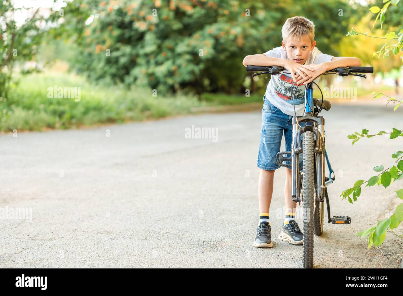 Müder Junge in T-Shirt und Jeansshorts lehnt sich nach einer langen Radtour im Park auf sein Fahrrad und zeigt Erschöpfung und Zufriedenheit Stockfoto