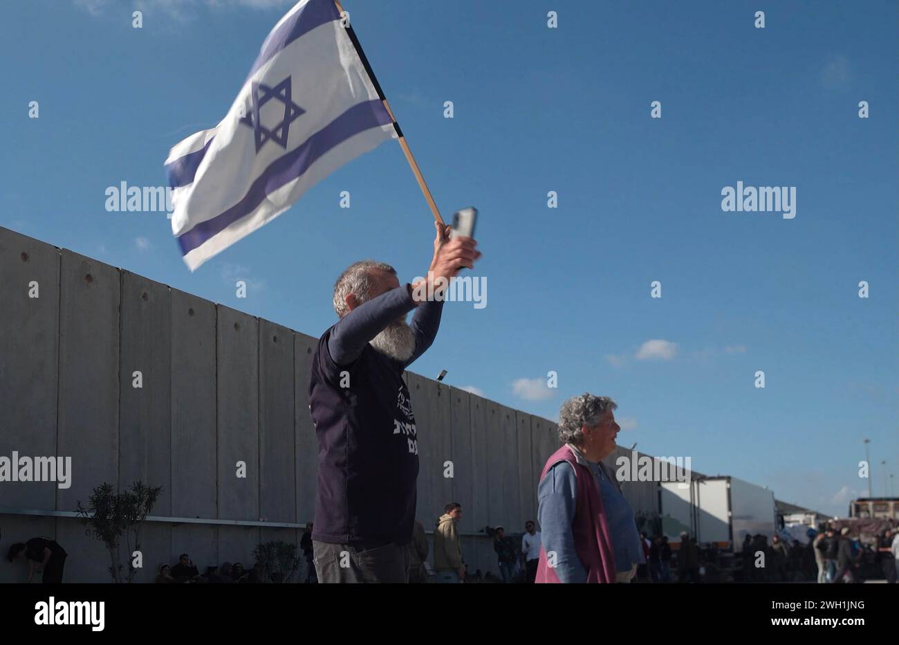 KEREM SHALOM, ISRAEL - 6. FEBRUAR: ein Demonstrant tanzt und hält eine israelische Flagge, während rechte israelische Demonstranten ägyptische Lastwagen blockieren, die humanitäre Hilfsgüter in den Gazastreifen auf der israelischen Seite des Grenzübergangs Karem Abu Salem transportieren, auch bekannt als Kerem-Schalom-Grenzübergang am 6. Februar 2024 in Kerem Schalom, Israel. Seit einigen Tagen blockieren israelische Siedler LKW, die humanitäre Hilfsgüter für Palästinenser im belagerten Gazastreifen transportieren. Stockfoto