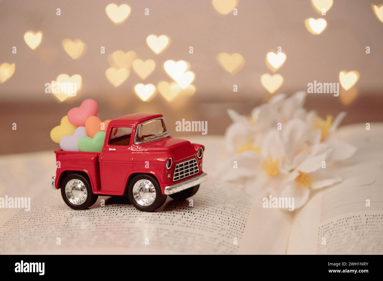Roter Truck trägt ein farbenfrohes Herz auf der Rückseite des Pickups, um Liebe zum Valentinstag zu liefern, auf einem verschwommenen Roman-Buch mit ausgebreiteter Seite, Herz-Bokeh Stockfoto