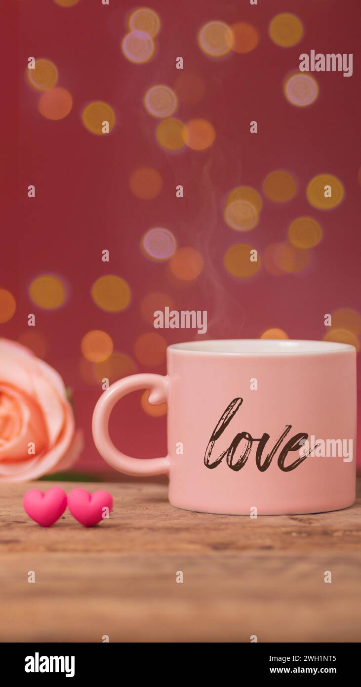 Heißgetränk mit Rauch in rosa Kaffeetasse, in der das Wort Love geschrieben wird, mit Rose und zwei kleinen Herzen nebeneinander auf Holztisch, Bokeh-Licht Stockfoto