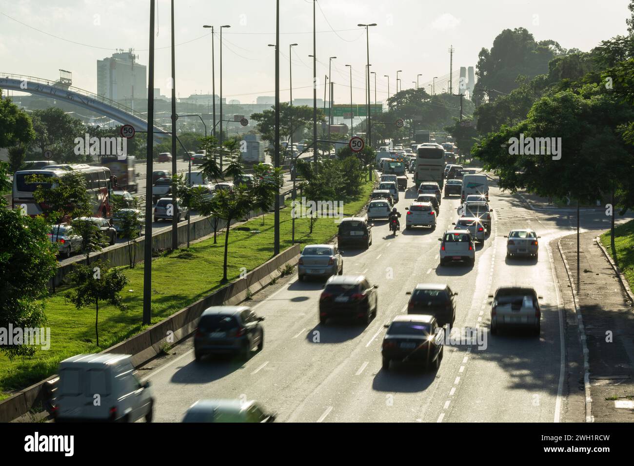 Tagsüber Blick auf eine geschäftige Straße in einer großen Stadt mit Autos, Bussen und Lastwagen. Viele Laternenpfosten, Bäume und Hintergrundbeleuchtung Stockfoto