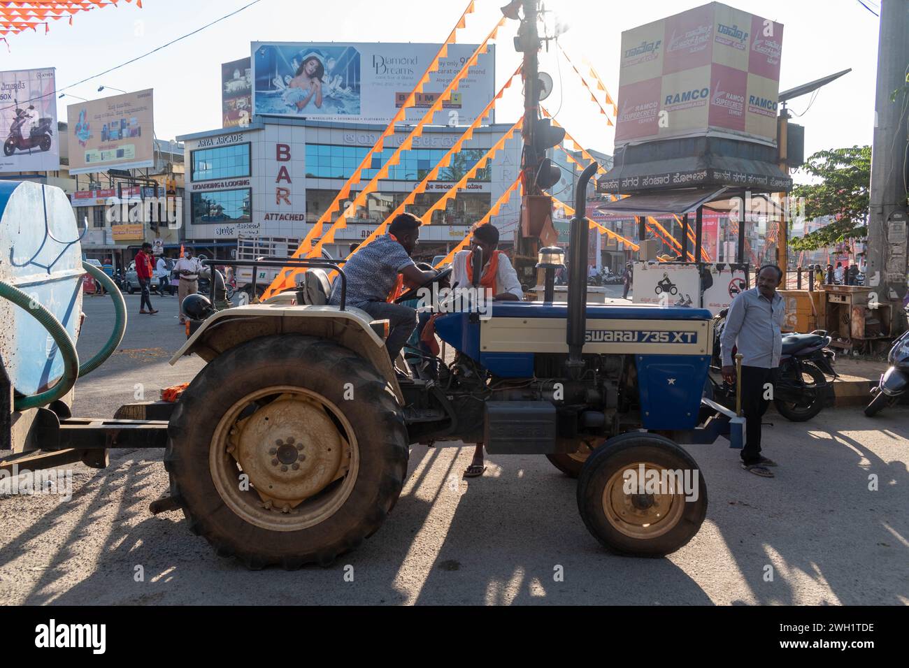 Hassan, Karnataka, Indien – 10. Januar 2023: Eine Gruppe von Männern wird auf einem Traktor gesehen, während sie durch eine geschäftige Straßenkreuzung in fahren Stockfoto