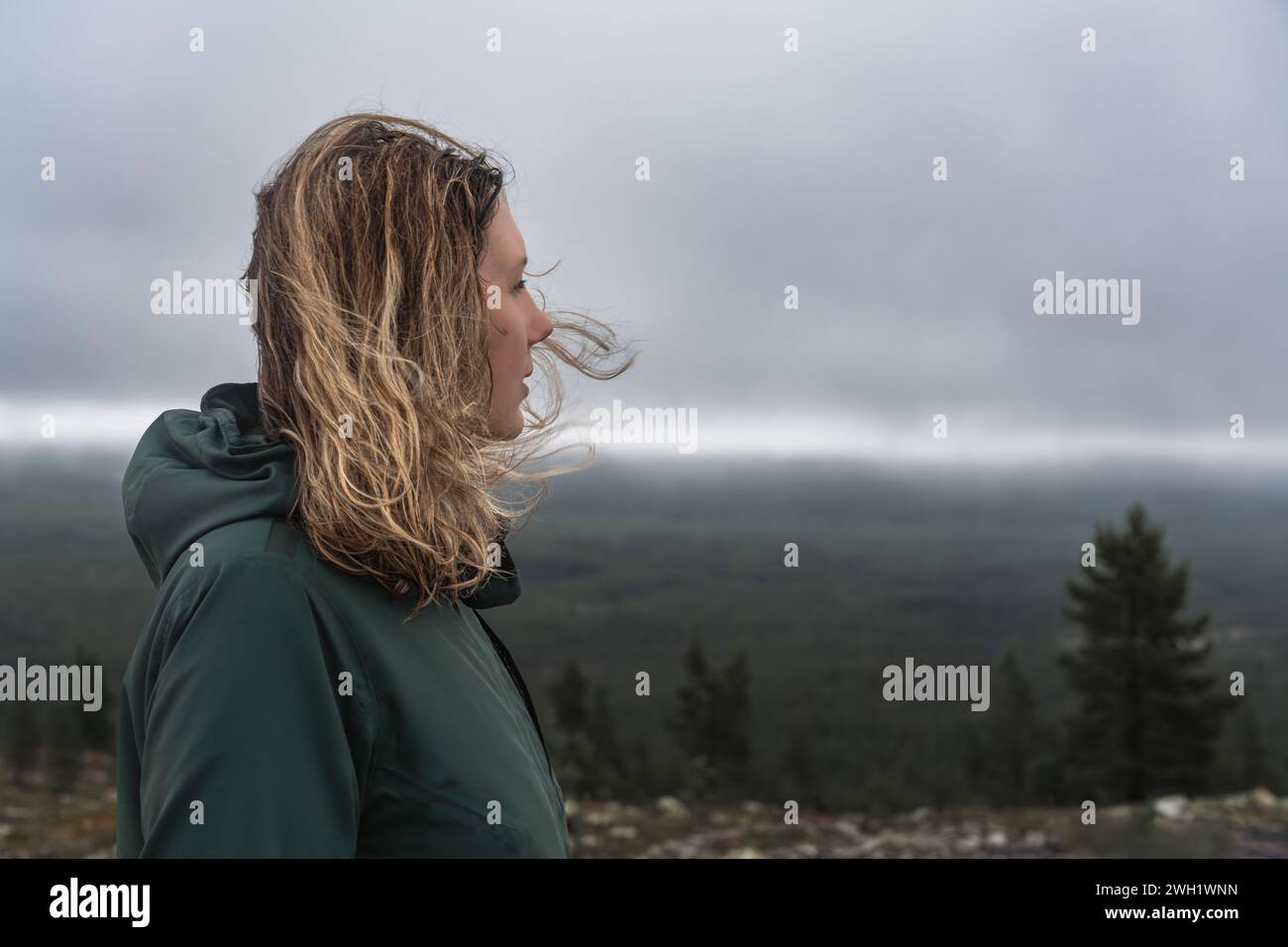 Seitenansicht einer Frau mit blonden Haaren, die in der nordischen Natur im Wind wehen, im Kopf, der über den Wald und die stürmischen Wolken im Hintergrund blickt Stockfoto