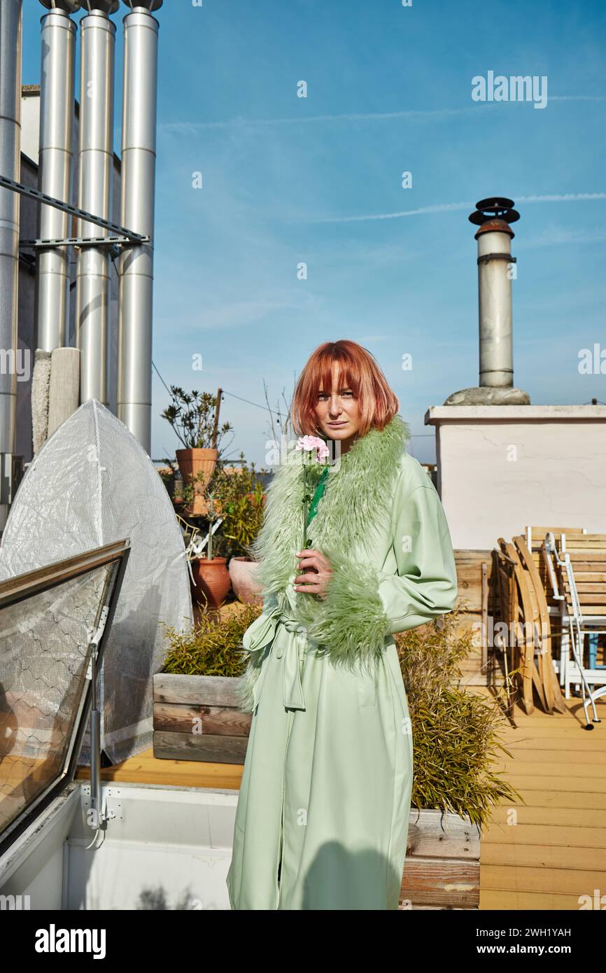 Trendige Frau in stilvoller Kleidung, die Blumen hält und auf der Dachterrasse in Wien in die Kamera blickt Stockfoto