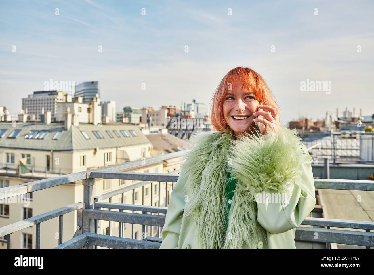 Lächelnde, stilvolle Frau, die auf dem Smartphone spricht und auf der Dachterrasse in Wien wegblickt Stockfoto