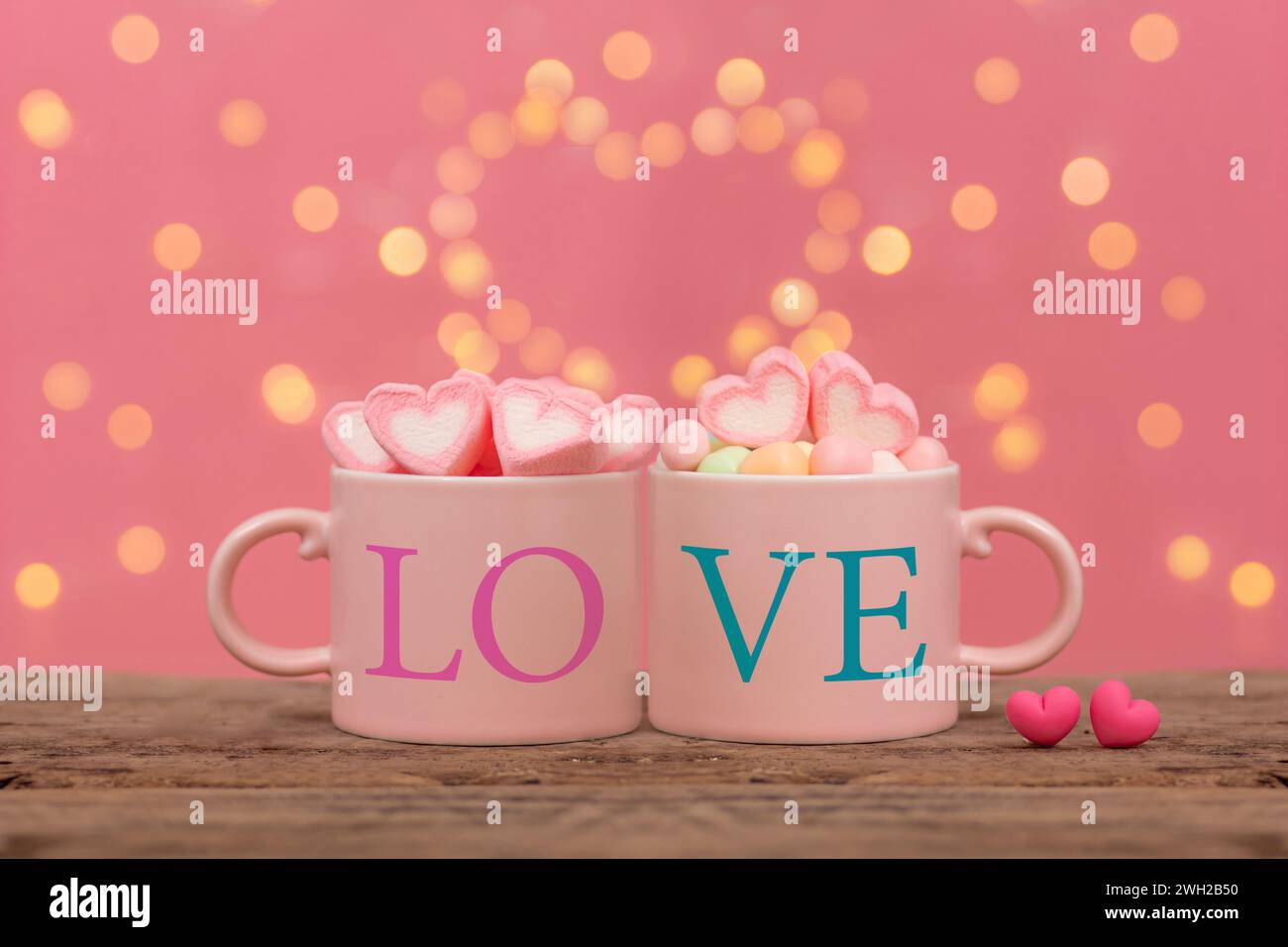 Ein paar pinkfarbene Kaffeetassen, auf denen das Wort Love steht, ein Marshmallow in Herzform, mit zwei kleinen Herzen nebeneinander auf einem Holztisch Stockfoto