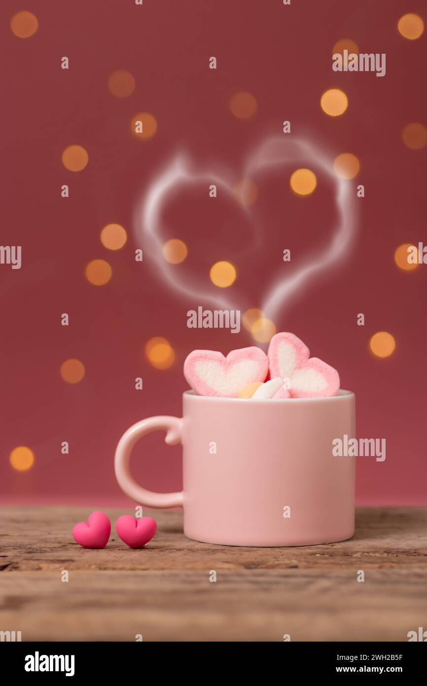 Heißgetränk mit herzförmigem Rauch auf rosafarbener Kaffeetasse mit herzförmigem Marshmallow oben, mit zwei kleinen Herzen nebeneinander auf Holztisch Stockfoto