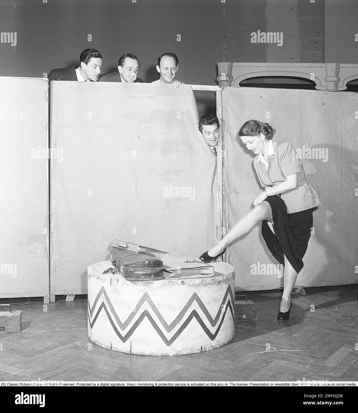 Hinter den Kulissen des Theaters in den 1950er Jahren Eine junge Frau steht und stellt die Strumpfhose an einem Bein ein, zeigt so mehr als sonst ihre schönen Beine. Es ist ungewiss, ob sie weiß, dass vier Männer stehen und auf sie schauen, während sie dies tut. 1950. Kristoffersson Ref. AY56-11 Stockfoto