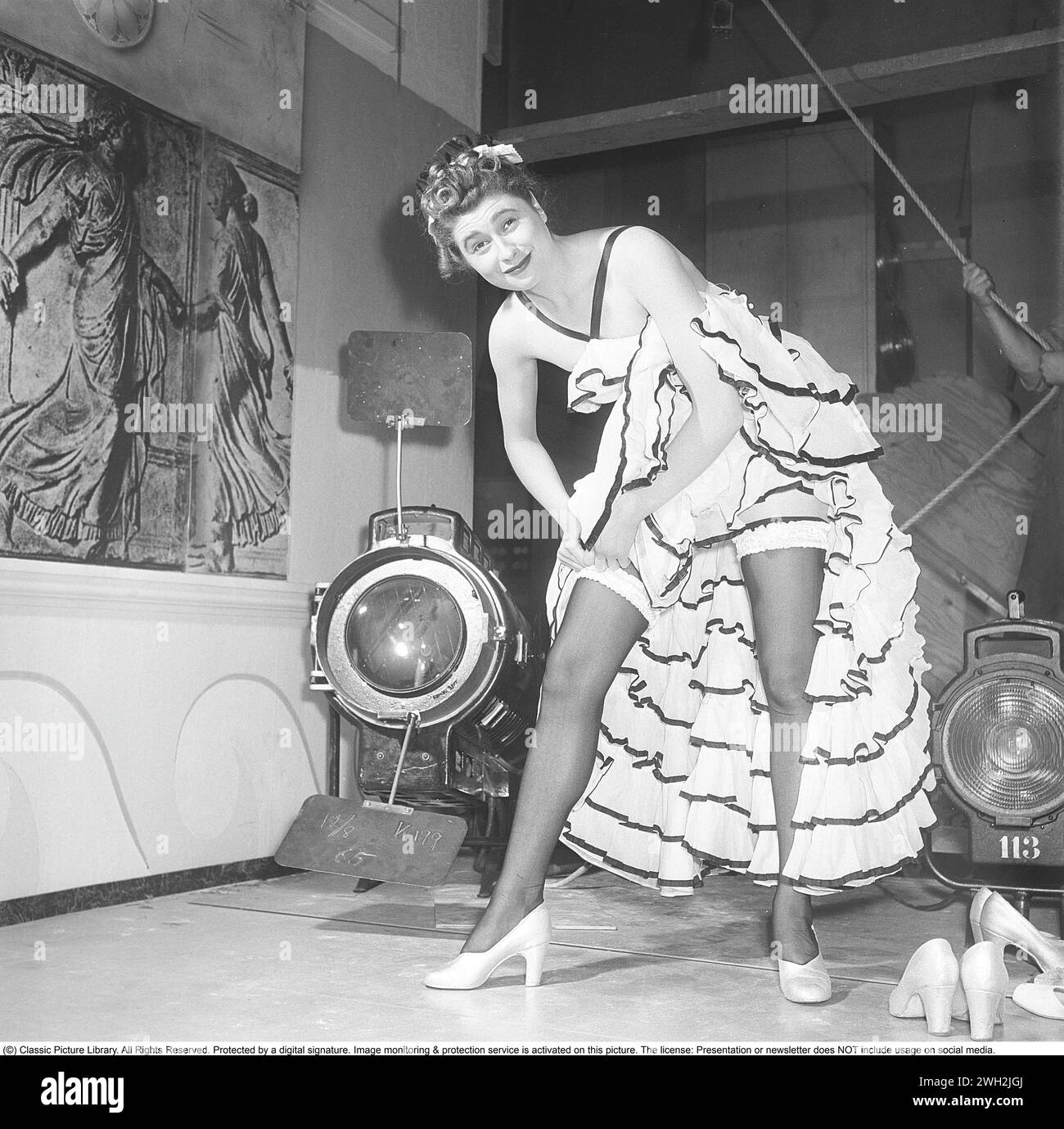 Hinter den Kulissen des Theaters in den 1950er Jahren Eine junge Frau aus einem Theaterensemble steht in ihrer Bühnenkleidung und hat ihren Rock hochgehoben, damit ihre Beine vollständig sichtbar sind. Die hohen Nylonstrümpfe werden an Strumpfhaltern befestigt. 1950. Kristoffersson Ref. AY64-10 Stockfoto