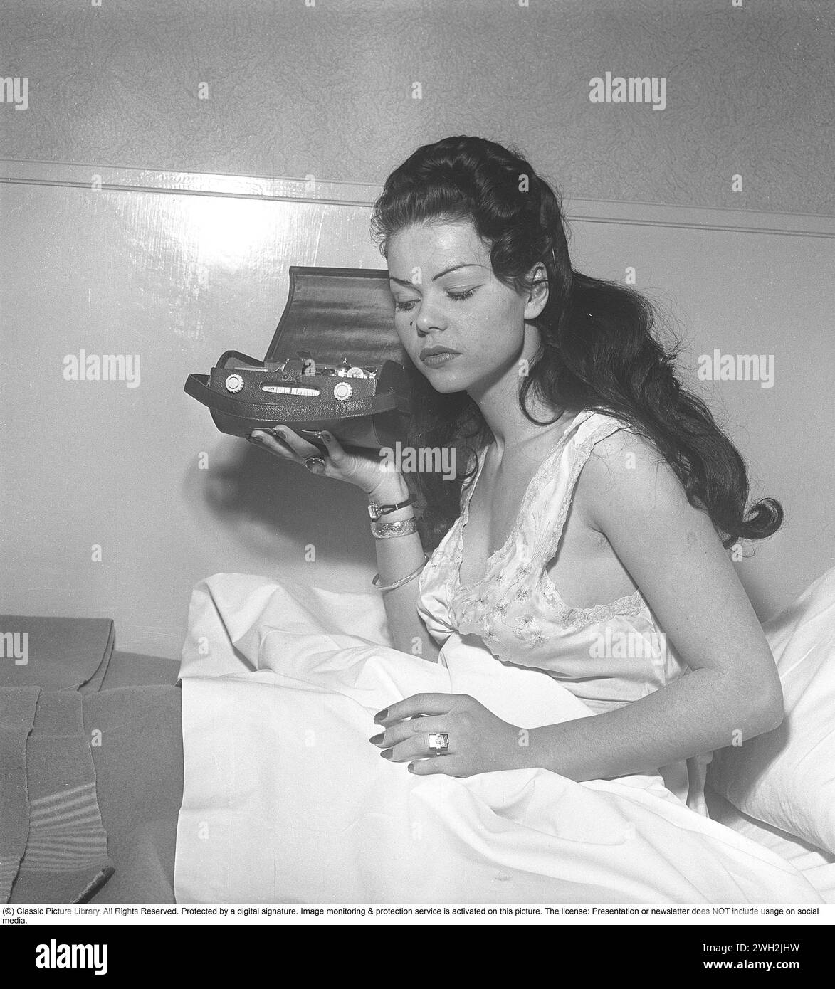 Radio in den 1950ern Eine junge dunkelhaarige Frau im Nachthemd sitzt mit ihrem tragbaren Transistorradio im Bett. Ein ungerades Modell, bei dem das äußere Gehäuse geöffnet und das Innere des Funkgeräts mit seinen Transistoren und Komponenten deutlich sichtbar ist. Schweden 1951. Kristoffersson Ref. BB29-6 Stockfoto