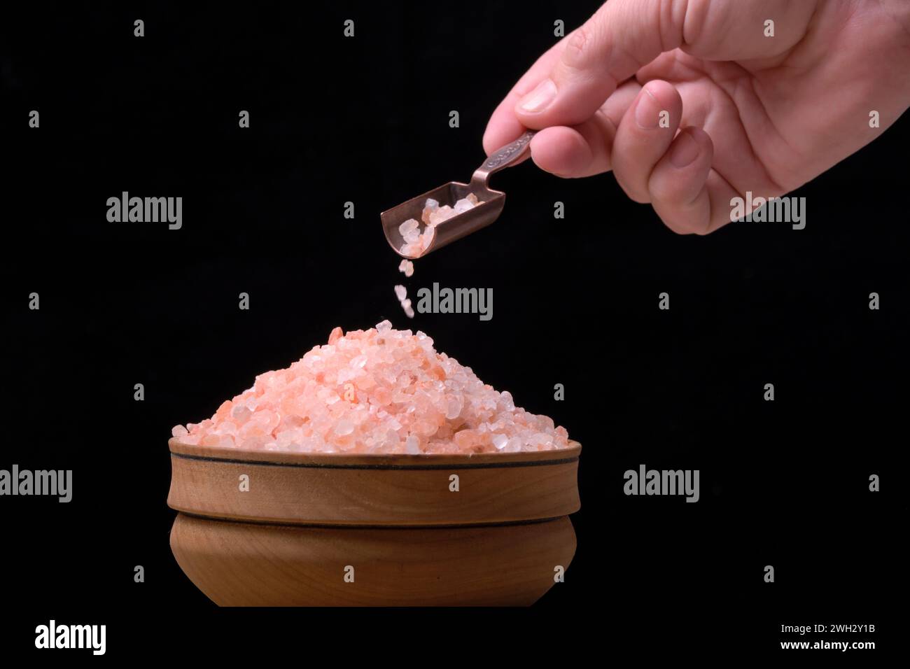 In seiner Hand gießt der Gewürzlöffel rosa Salz in den Becher auf schwarzem Hintergrund Stockfoto