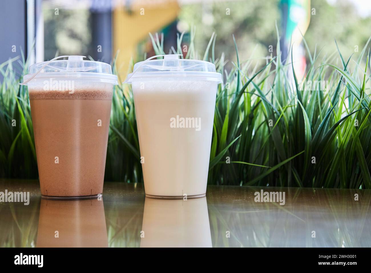 Zwei klare Plastikgläser mit Deckel gefüllt mit Schokoladenvanilla-Milchshakes auf dem Tisch, grünes Gras. Leeres Modell, Hintergrundgetränk-Café. Mitnehmen Stockfoto