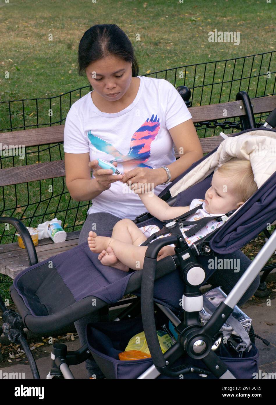 Ein Kindermädchen schneidet die Fingernägel eines Kindes mit einem etwas, das wie ein spezieller Clipper für Kinder aussieht. Er scheint unglücklich zu sein. Im Washington Square Park in Manhattan. Stockfoto