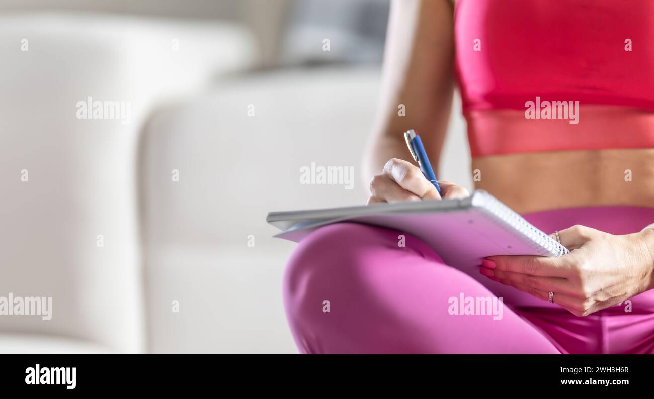 Sportliche Frau sitzt auf einer Matte und schreibt zukünftige Trainingspläne auf, um sportliche Ergebnisse zu erzielen. Stockfoto