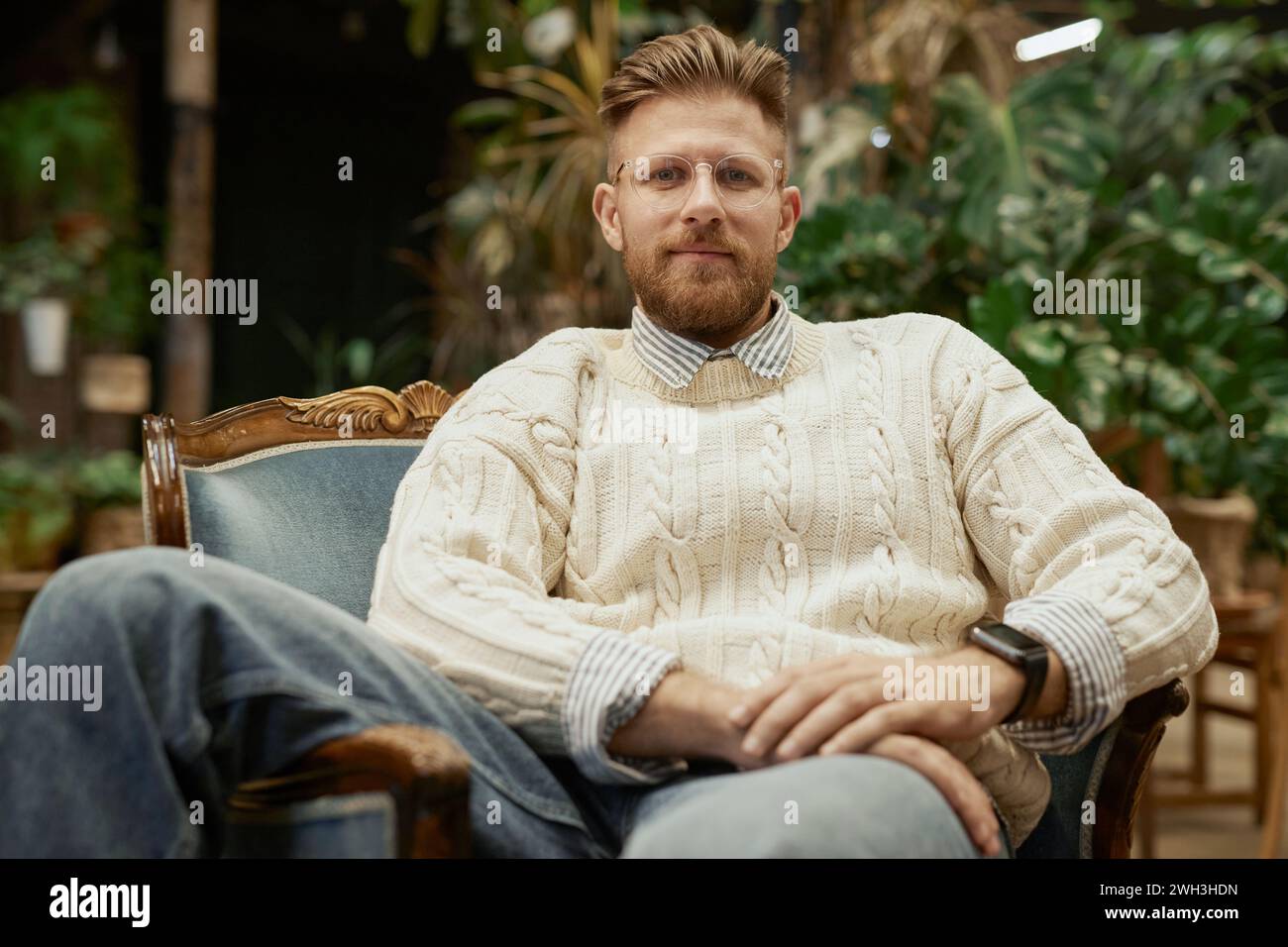 Porträt eines bärtigen jungen Mannes mit Brille und lässigem Pullover, der in einem Vintage-Stuhl liegt und selbstbewusst in die Kamera blickt Stockfoto